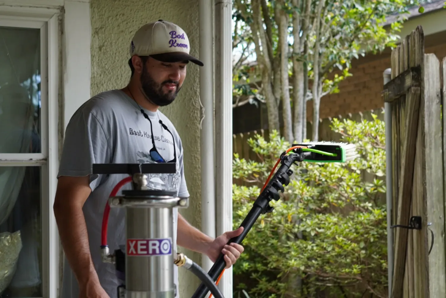 Boot Krewe Cleaners technician in a branded cap and grey tee standing beside the pure-water filter tank, holding the brush head of his pole
