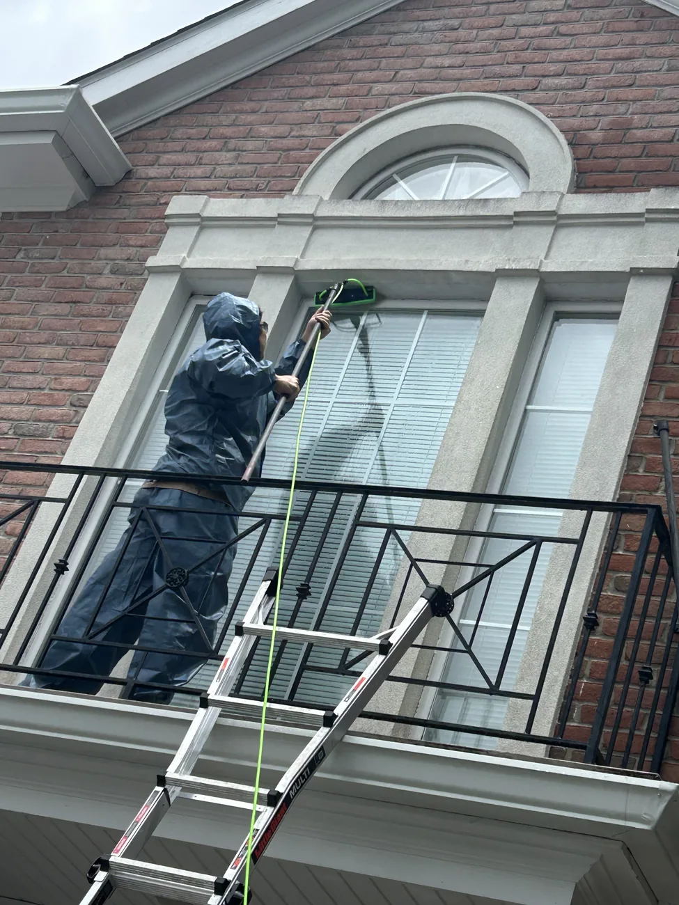 Technician in wet weather gear on a balcony cleaning an arched upper window of a brick house