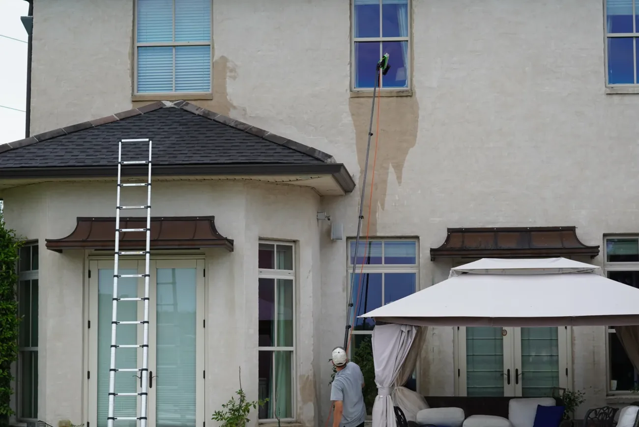 Cream stucco two-storey house with a ladder against the lower roof and a pole reaching a second-storey window, water running down the wall