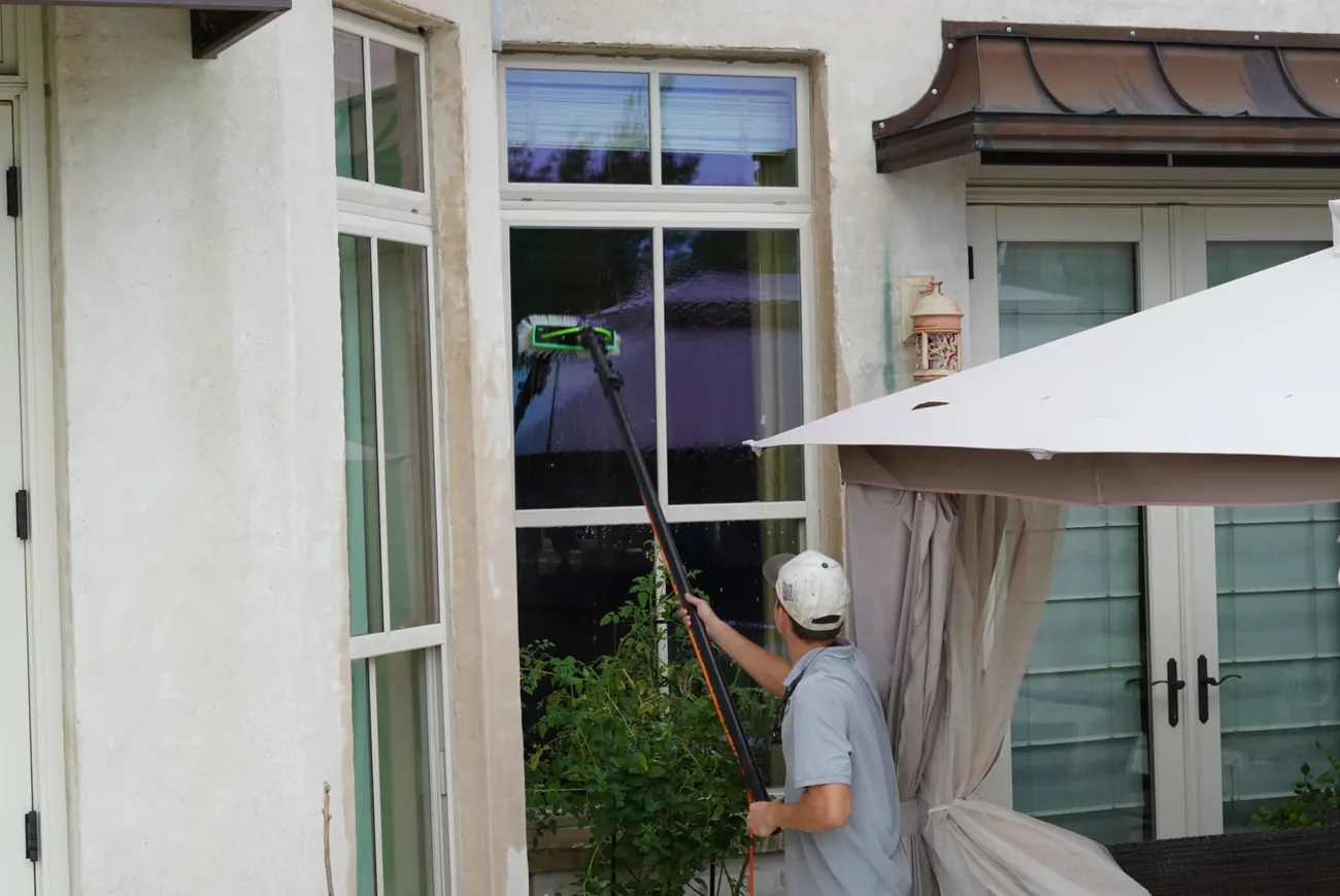 Technician reaching a tall upper window with a water fed pole and green brush head, water running down the glass